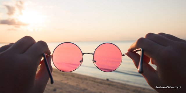 Różowe okulary trzymane w dłoniach na tle zachodu słońca nad wodą